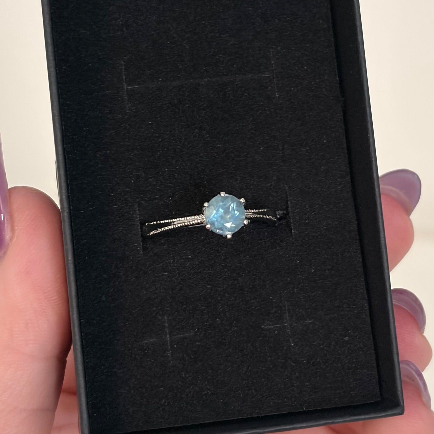 A silver ring with a round light blue gemstone is displayed inside an open black jewelry box, held by a hand with purple nail polish.